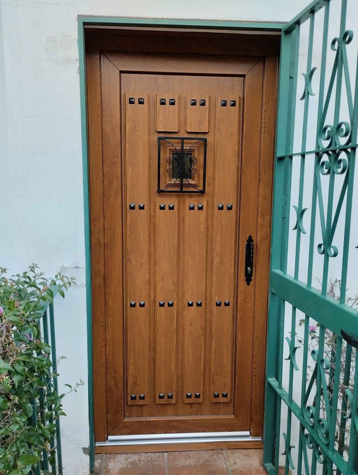 Puerta de entrada de madera con una pequeña ventana en una pared blanca, junto a una verja metálica verde y una planta en maceta.