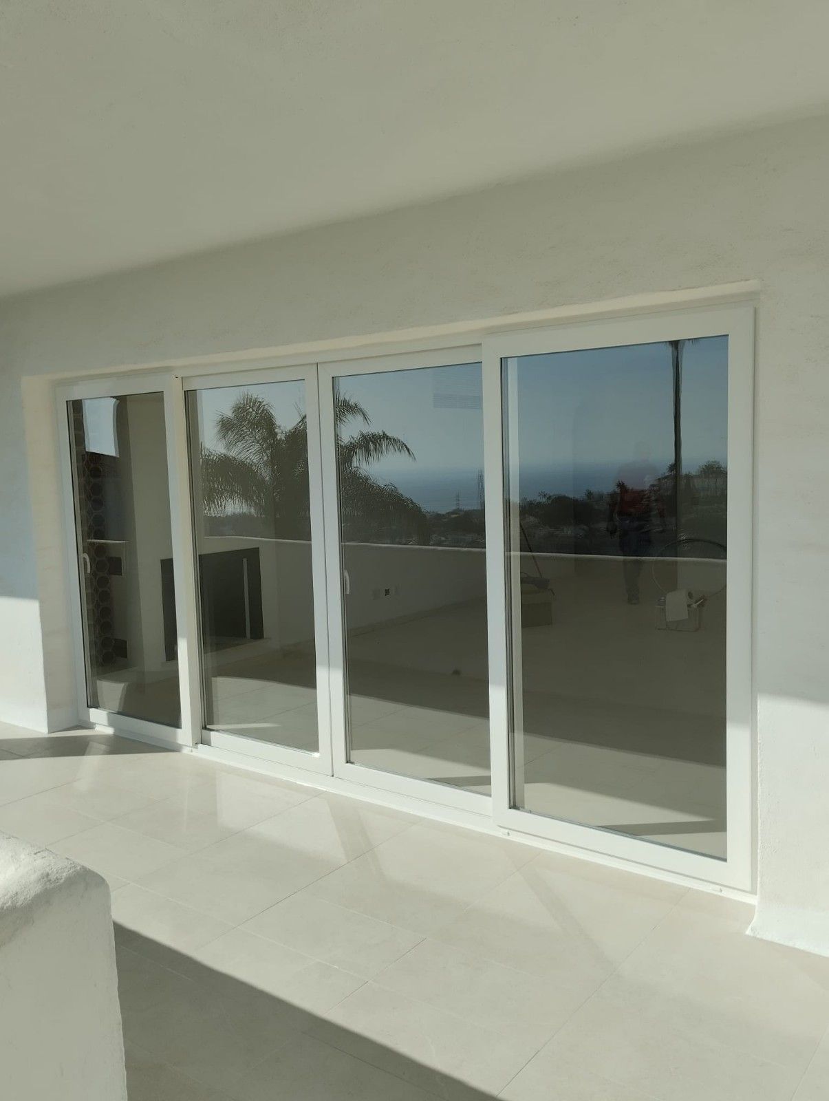 Patio blanco con grandes puertas corredizas de vidrio que reflejan las palmeras y la vista exterior.