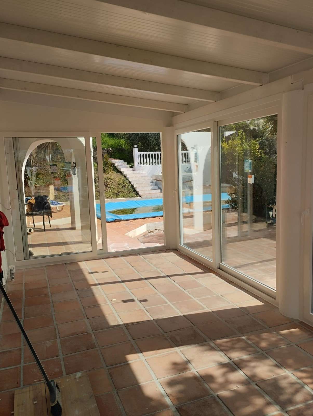 Patio cerrado y soleado con baldosas de terracota, vigas blancas y puertas de cristal que dan a una piscina y un jardín.
