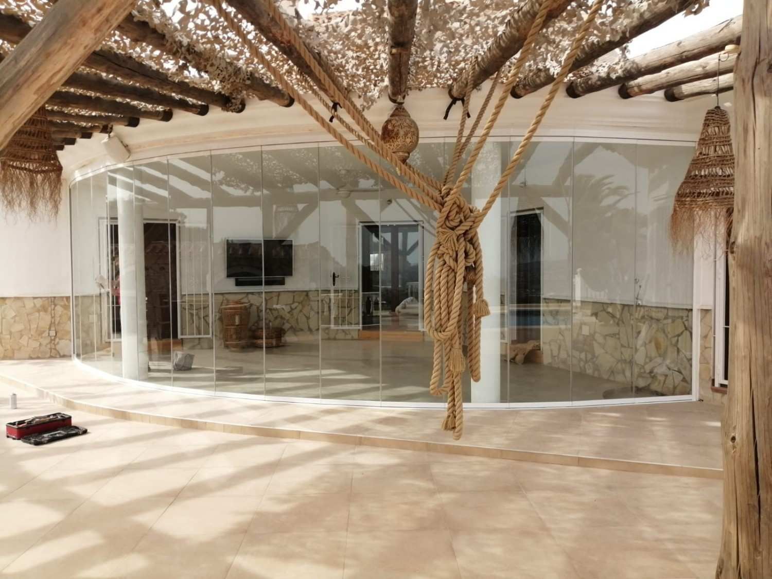 Espacio interior luminoso con pared de cristal, suelo de piedra y hojas de palmera colgantes.