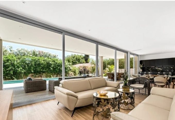 Sala de estar moderna con sofás beige, mesas de centro de cristal y ventanales que van del suelo al techo y dan a un patio con vegetación.