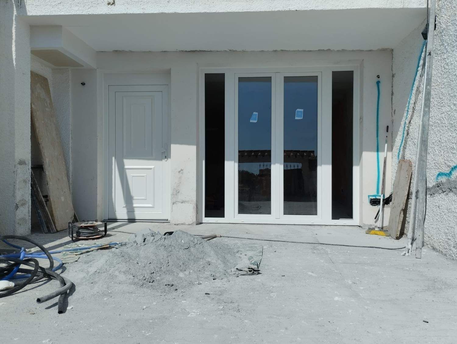 Exterior de una casa blanca con puertas dobles de cristal y una puerta individual en un patio de hormigón durante la construcción.