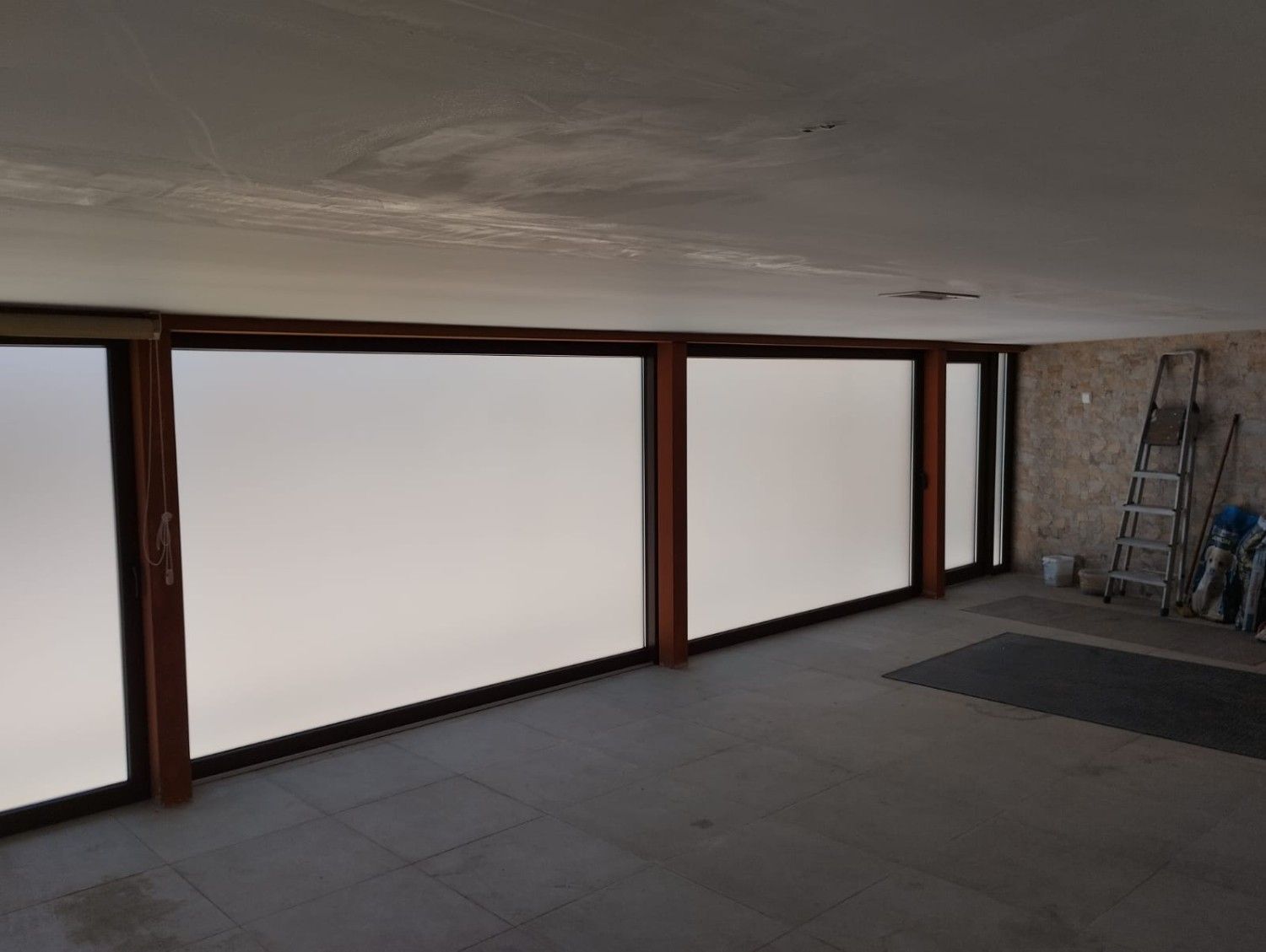 Habitación vacía con grandes ventanas esmeriladas, suelo oscuro y una escalera en la esquina.