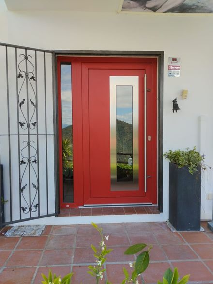 Puerta principal roja con paneles de vidrio estrechos, junto a una verja de seguridad metálica y una planta en maceta en el porche.