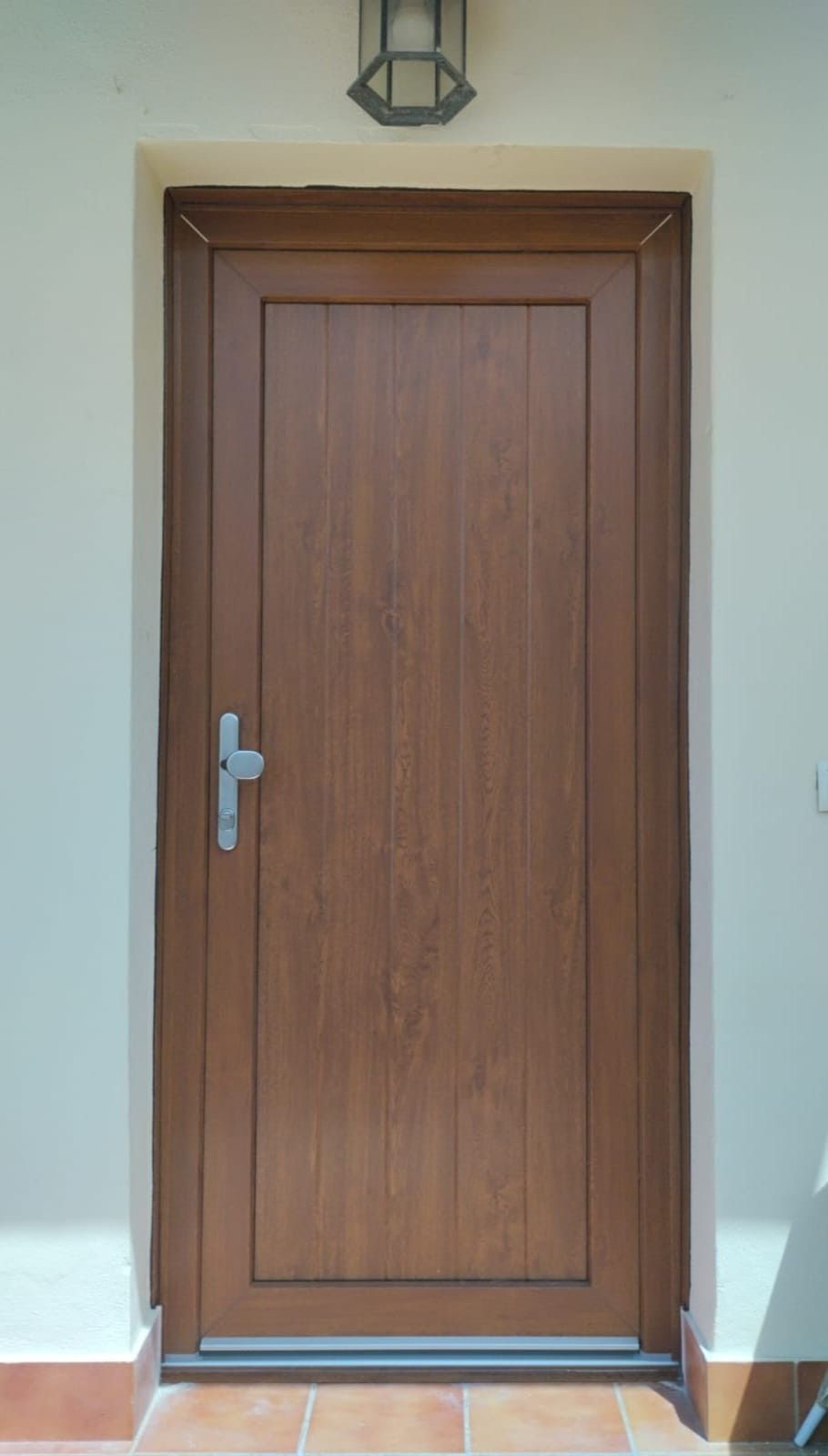 Puerta principal de madera cerrada con tirador plateado debajo de una lámpara de pared.