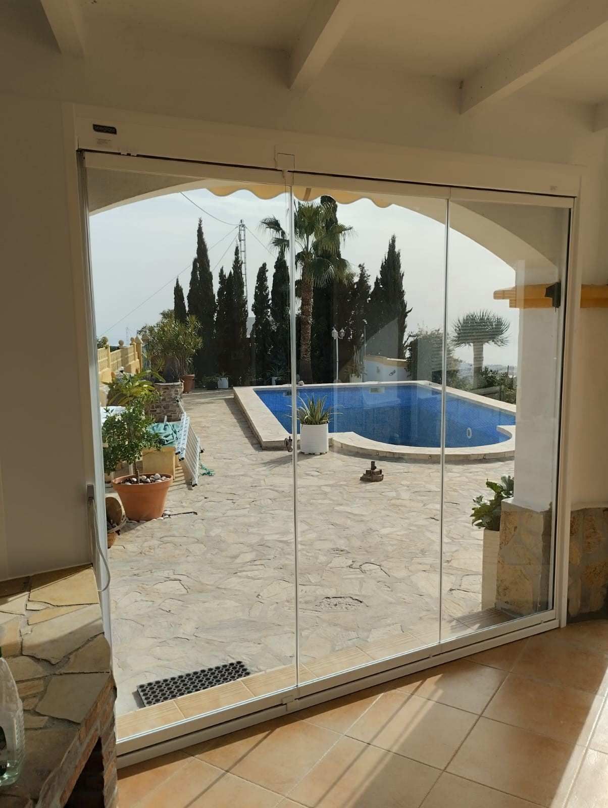 Unas puertas corredizas de cristal dan acceso a un patio soleado con una piscina azul, plantas en macetas y árboles al fondo.