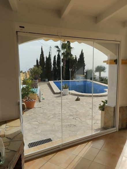 Unas puertas corredizas de cristal dan acceso a un patio soleado con una piscina azul, plantas en macetas y árboles al fondo.