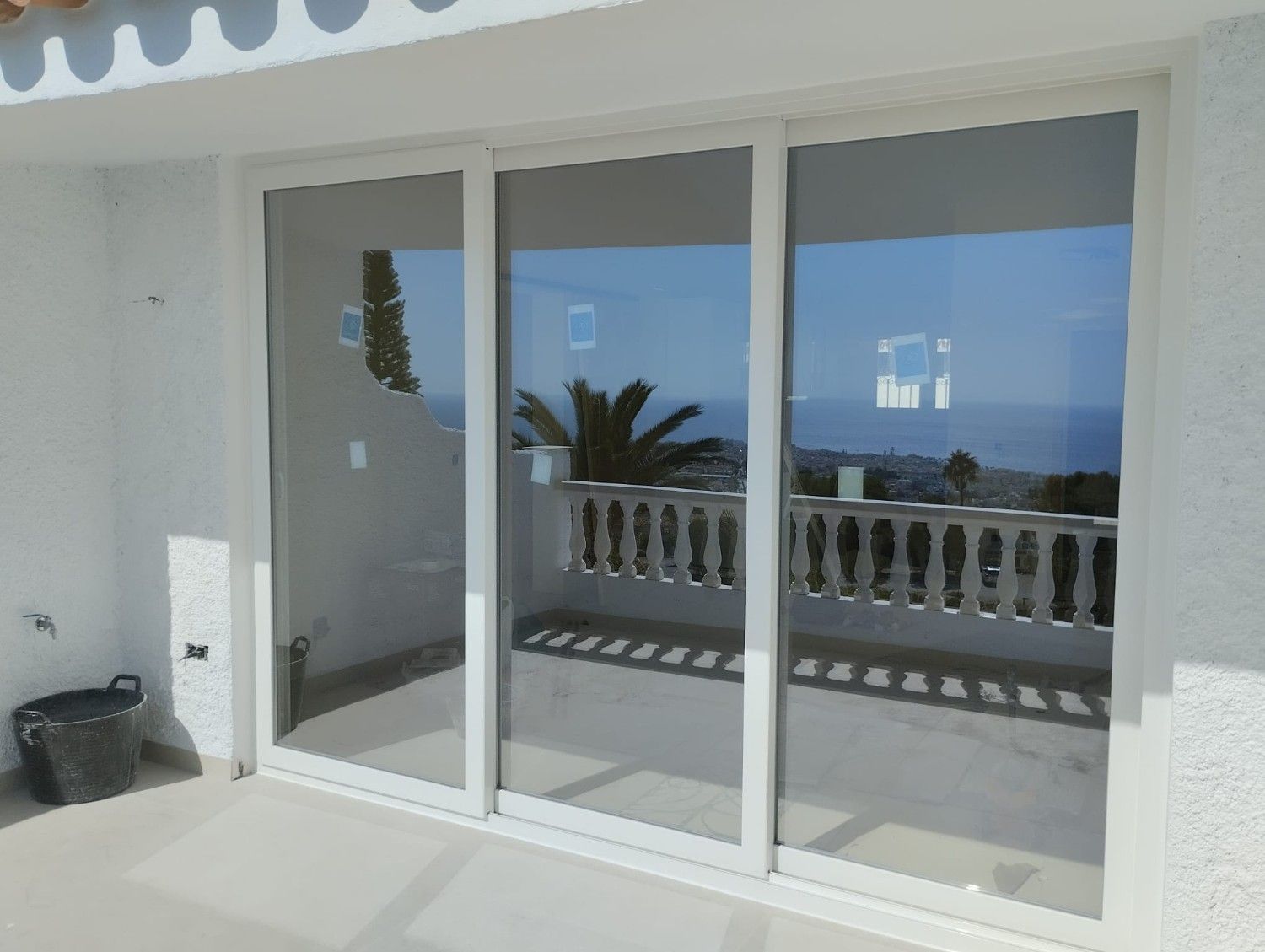 Puertas corredizas de cristal que dan a un balcón soleado con vistas al océano, barandilla blanca y cielo azul.