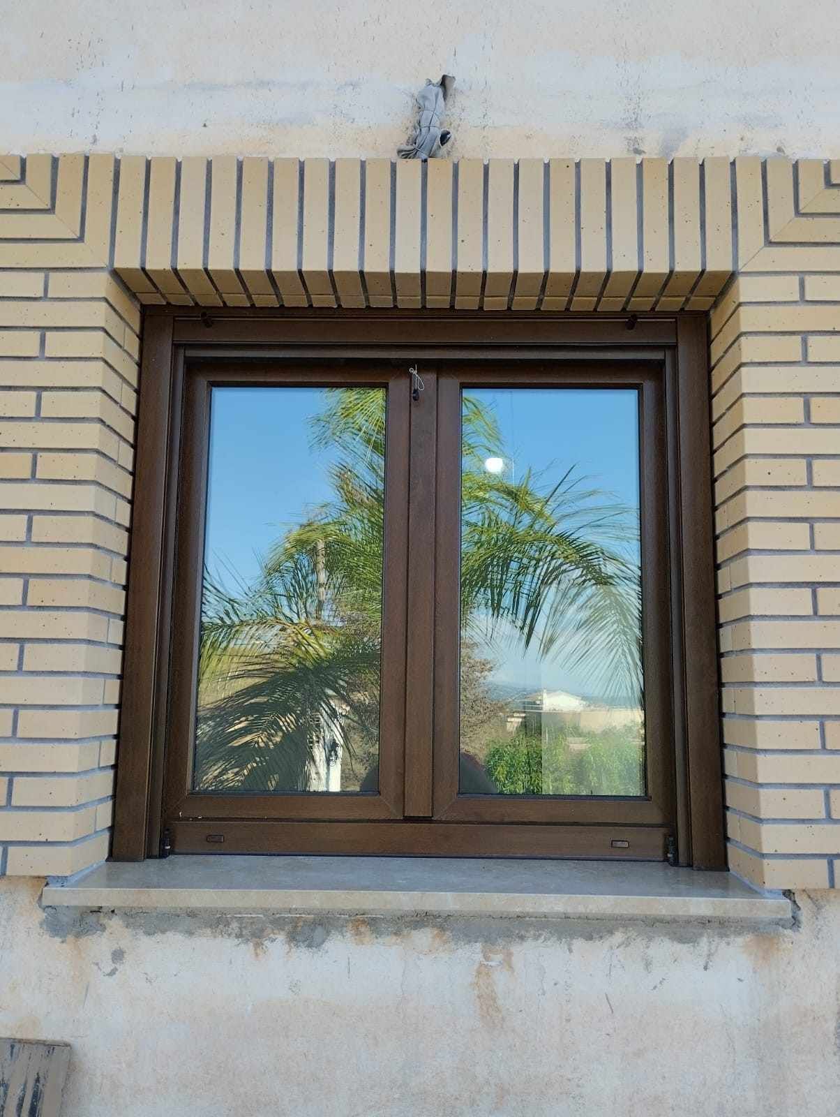 Ventana con marco marrón en una pared de ladrillo beige con una palmera reflejada en el cristal.