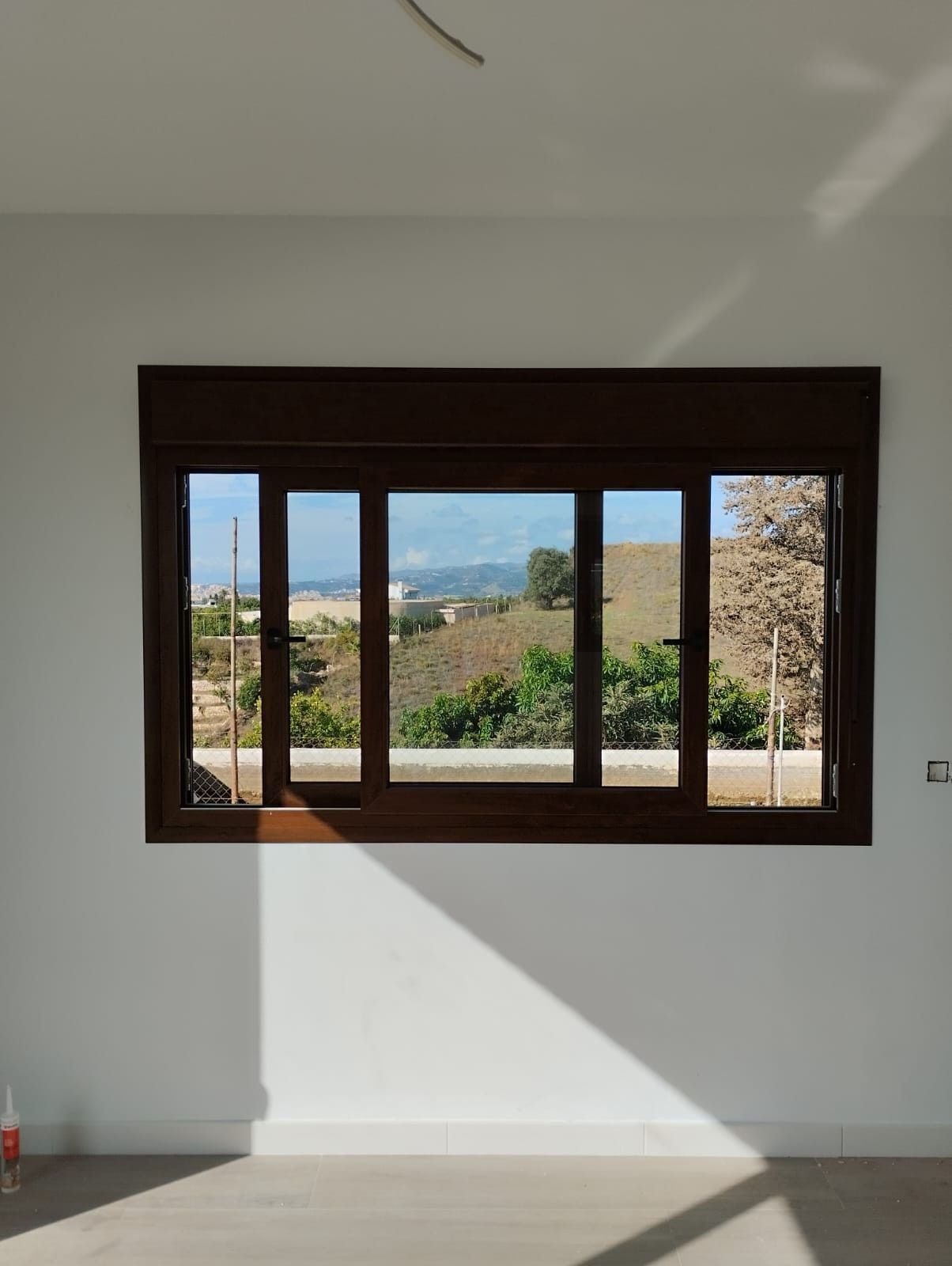 Pared blanca con una ventana de madera oscura que enmarca una vista exterior soleada de colinas y árboles.