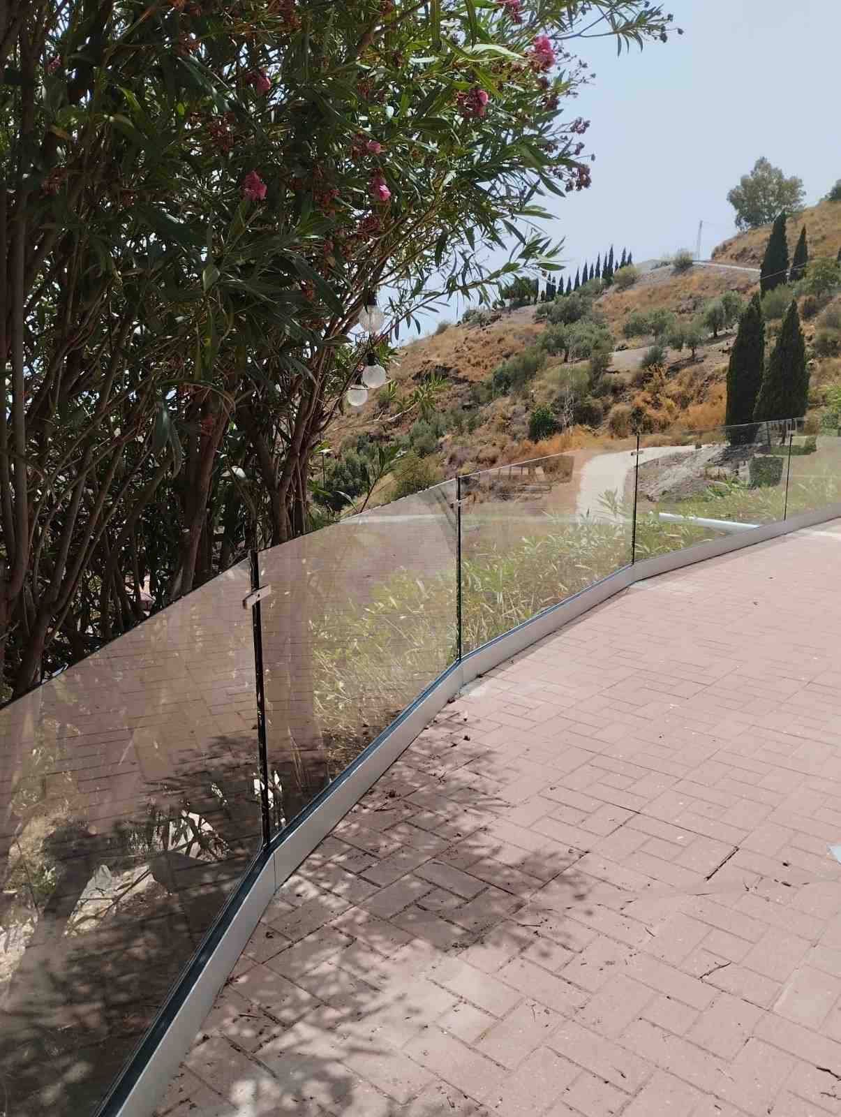 Sendero curvo de ladrillos junto a una valla, árboles y una ladera seca bajo un cielo despejado.