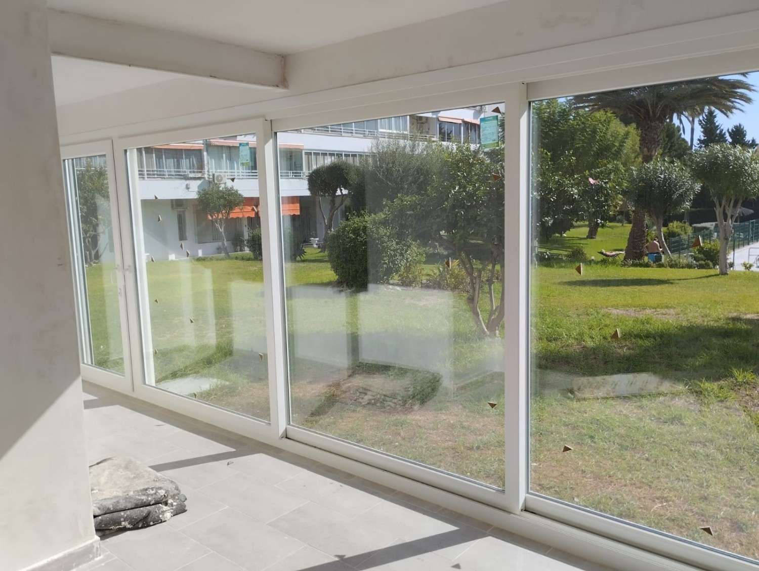 Habitación vacía y soleada con ventanales que van del suelo al techo y dan a un patio cubierto de césped.
