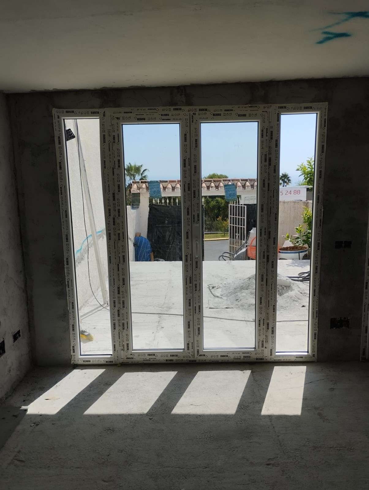 Habitación interior con altas puertas de cristal que dan a un balcón soleado, proyectando sombras de las ventanas sobre el suelo.