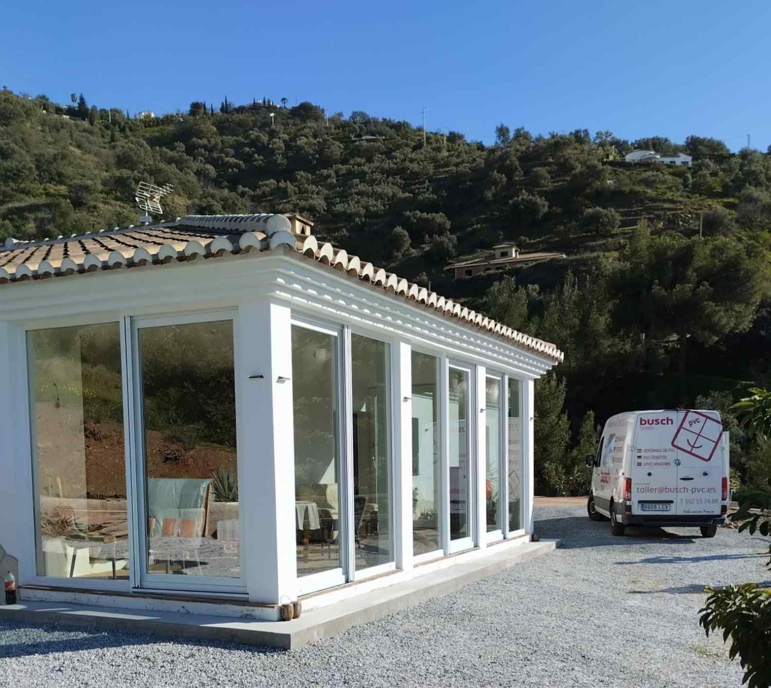 Un invernadero de cristal blanco en construcción en un terreno de grava con una furgoneta y una ladera boscosa detrás.