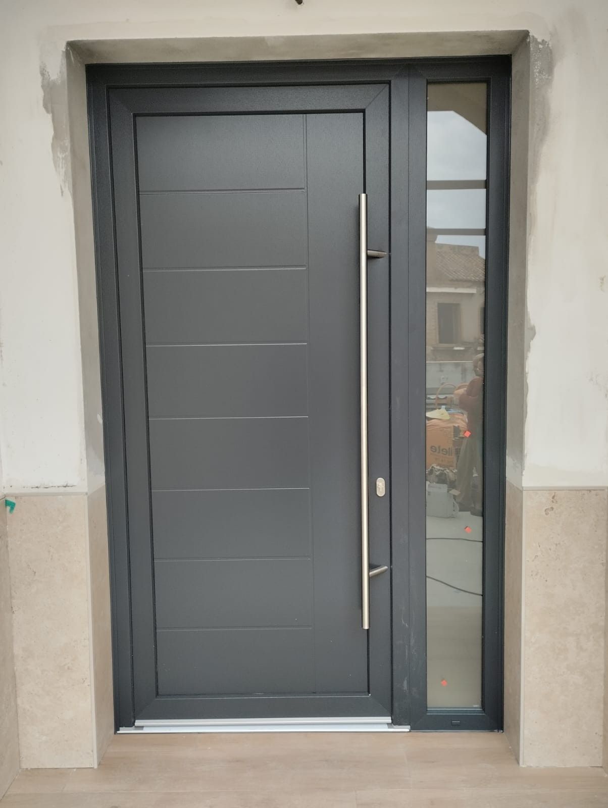 Puerta principal gris con un panel lateral de vidrio estrecho en una pared exterior blanca.