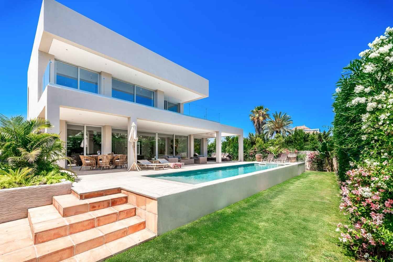 Casa blanca moderna con piscina, terraza y un exuberante césped verde bajo un cielo azul despejado.