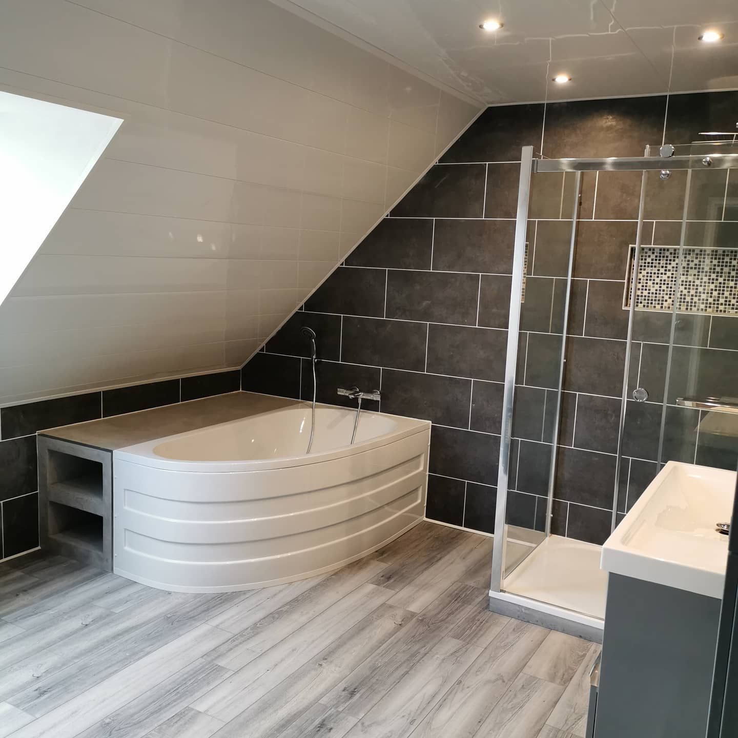 Salle de bains moderne avec plafond en pente, carrelage gris et blanc, baignoire incurvée, douche et meuble lavabo.