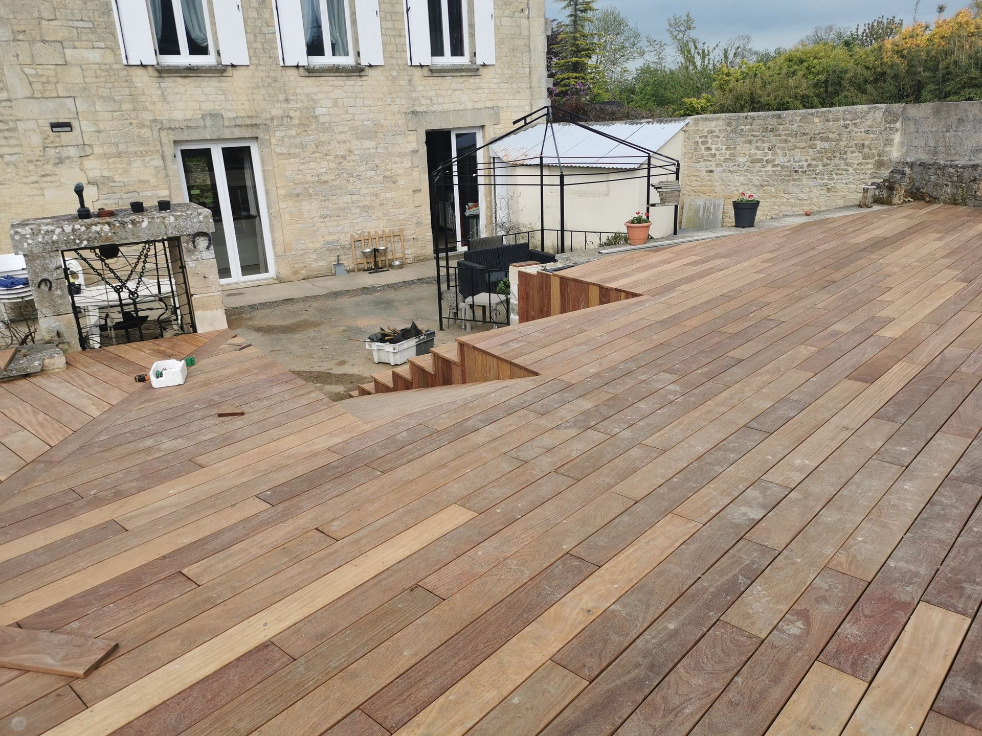 Terrasse en bois avec marches descendantes, à côté d'un bâtiment en pierre avec fenêtres et jardin.