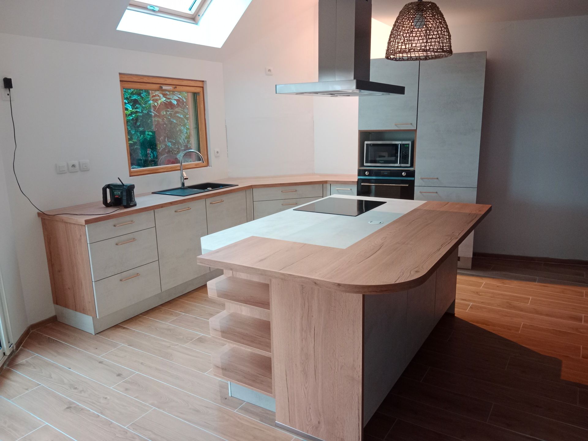 Cuisine moderne avec armoires en bois clair, îlot central et électroménager. Puits de lumière au-dessus de l'évier et plancher en bois.