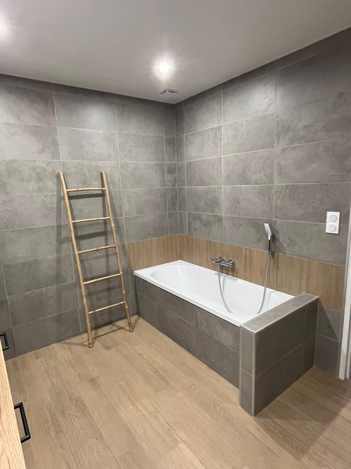 Salle de bains aux murs carrelés de gris, au sol en bois clair et équipée d'une baignoire. Une échelle en bois est appuyée contre le mur.