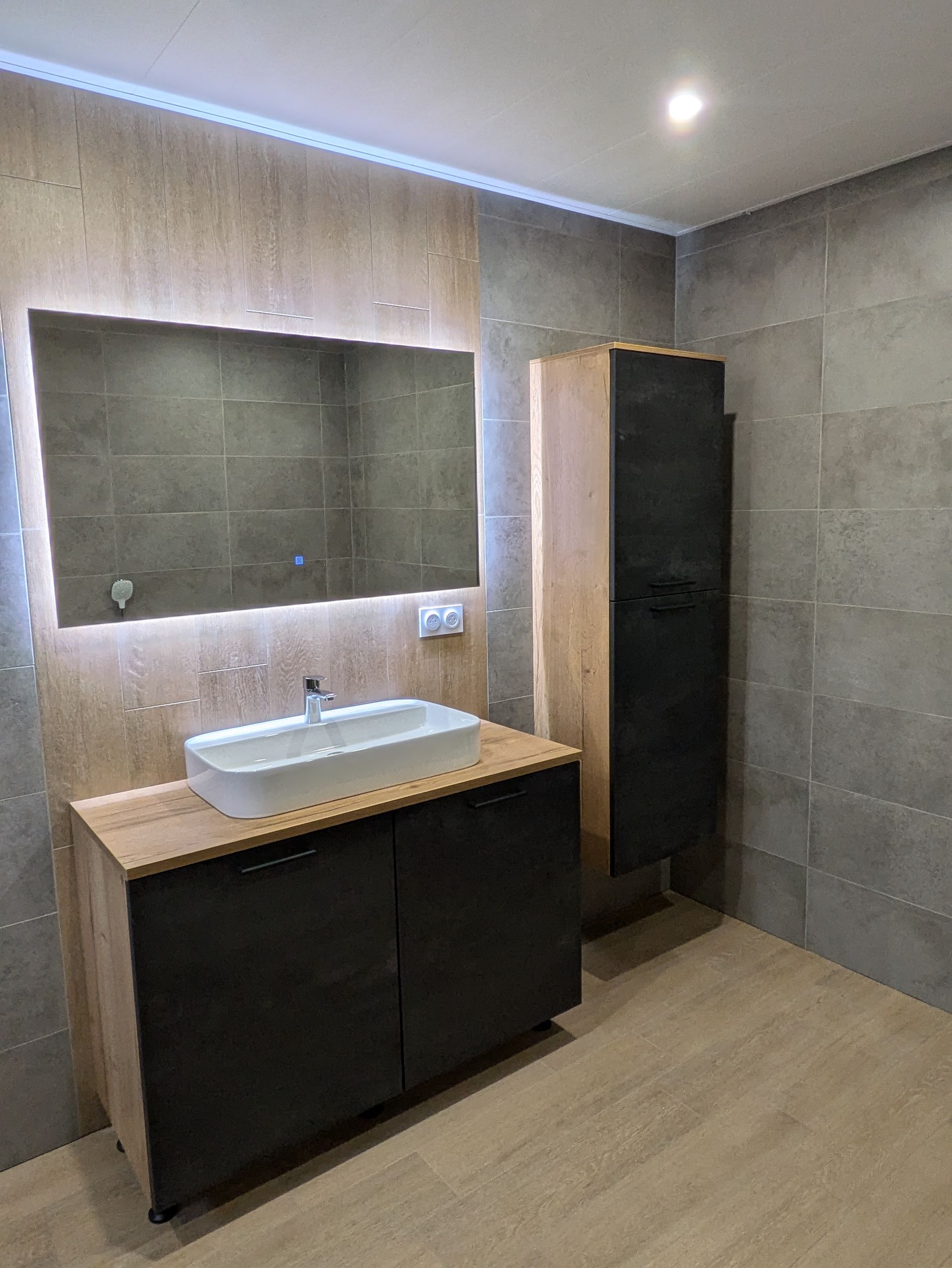 Salle de bains moderne avec meuble lavabo, miroir et armoire haute. Carrelage gris et touches de bois.