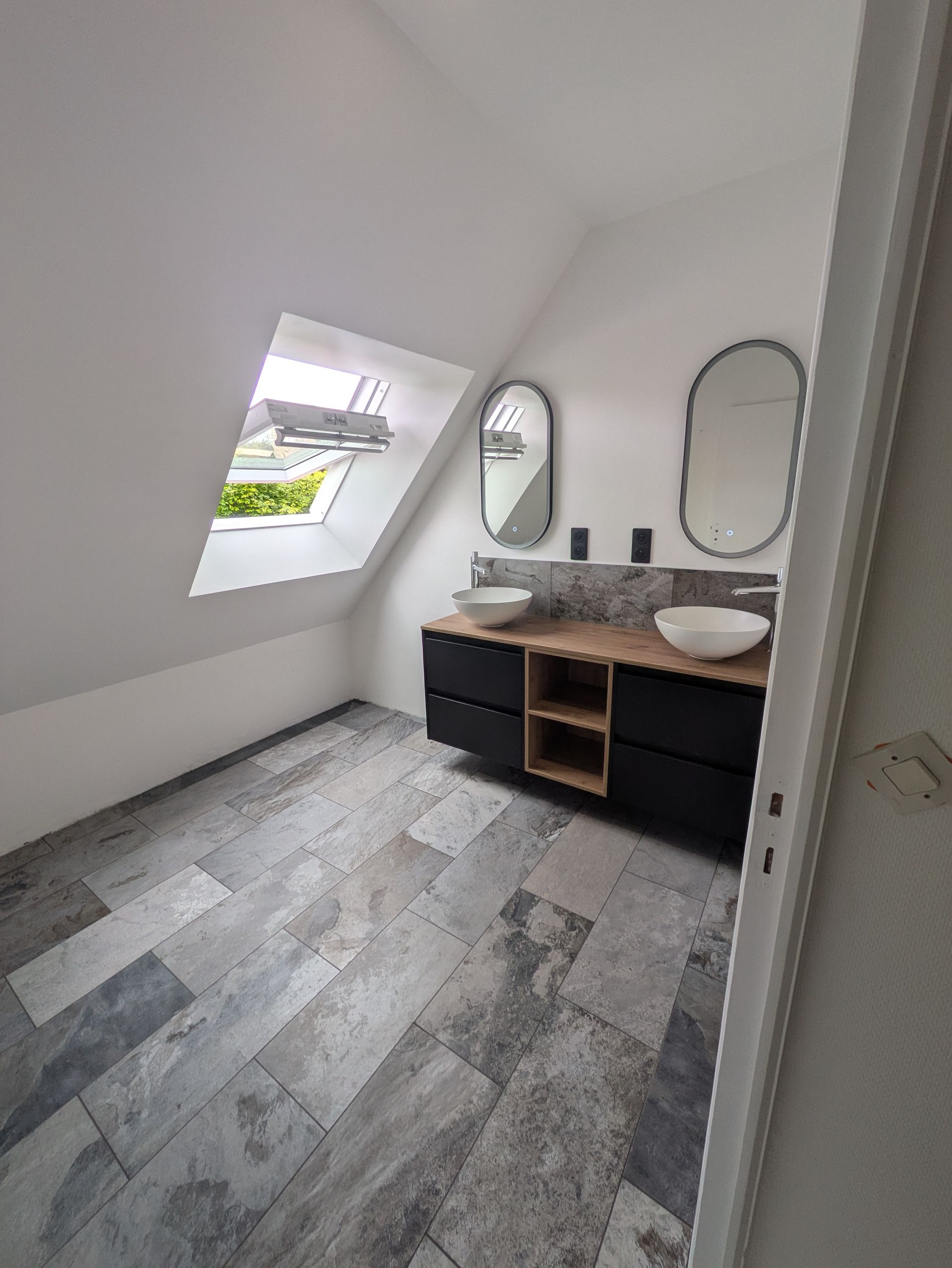 Salle de bains avec plafond en pente, puits de lumière, meuble-lavabo noir, deux miroirs ovales et sol carrelé gris.