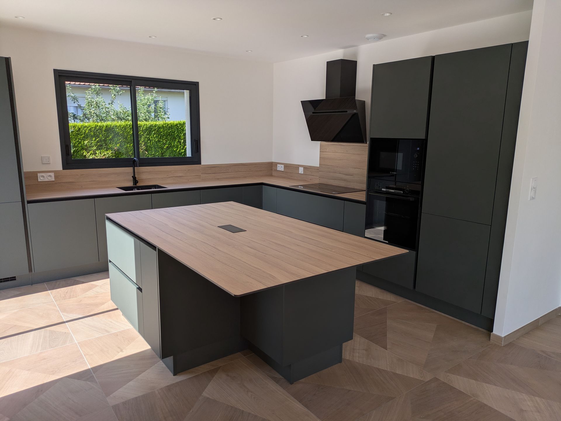 Cuisine moderne avec placards gris, plan de travail en bois, îlot central et fenêtre.