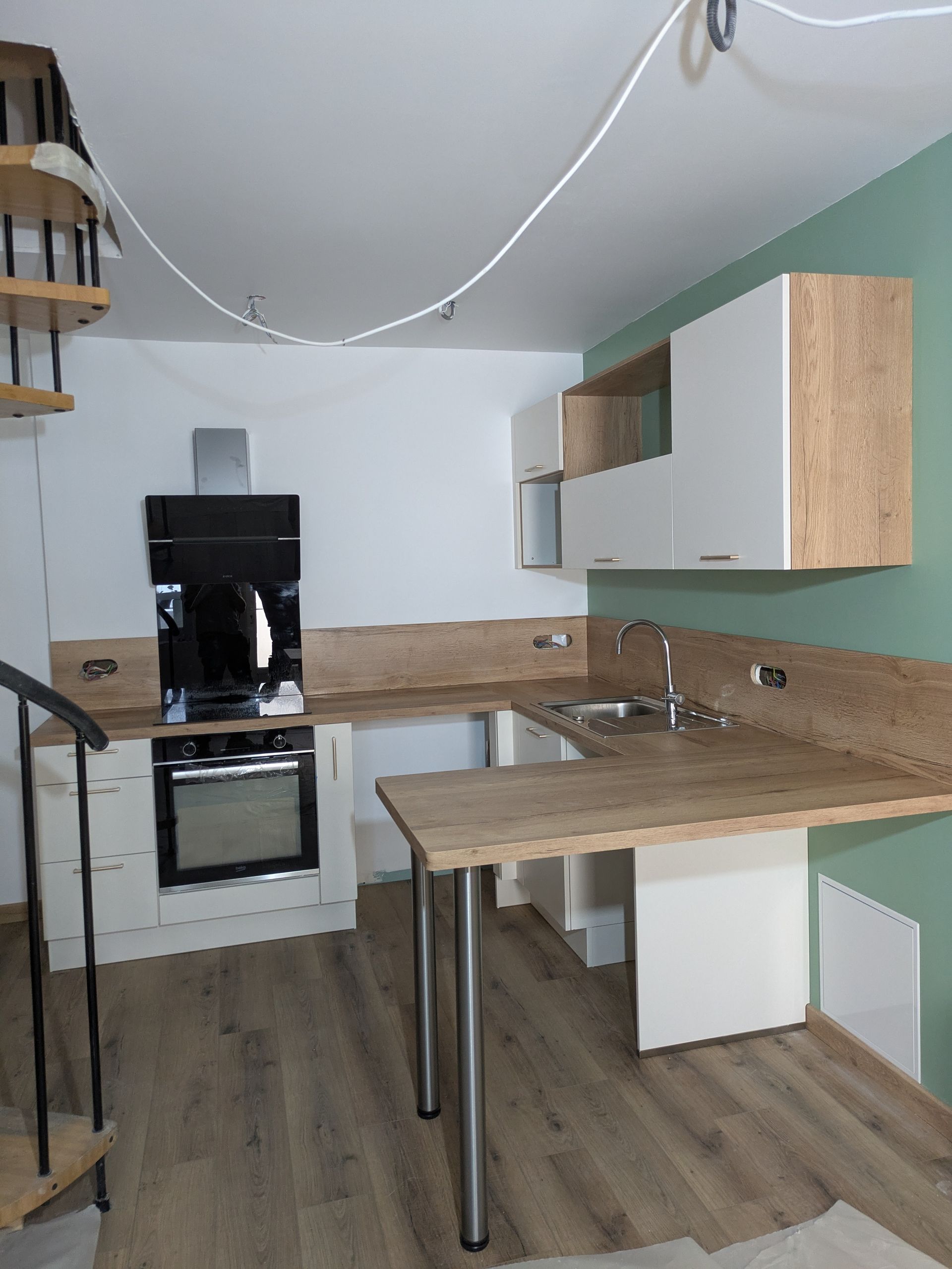 Petite cuisine avec placards blancs et bois, four et évier ; une petite table prolonge le plan de travail.