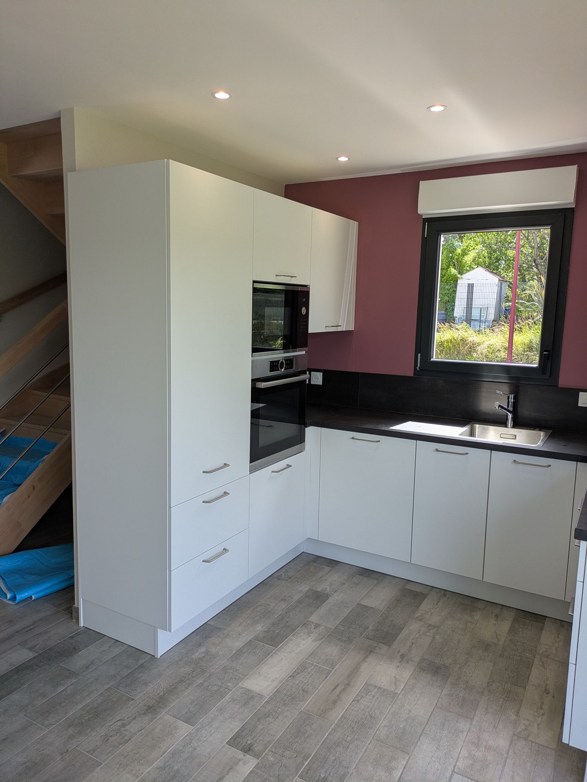 Cuisine moderne blanche avec comptoirs, four et micro-ondes foncés. Sol gris et fenêtre avec vue.