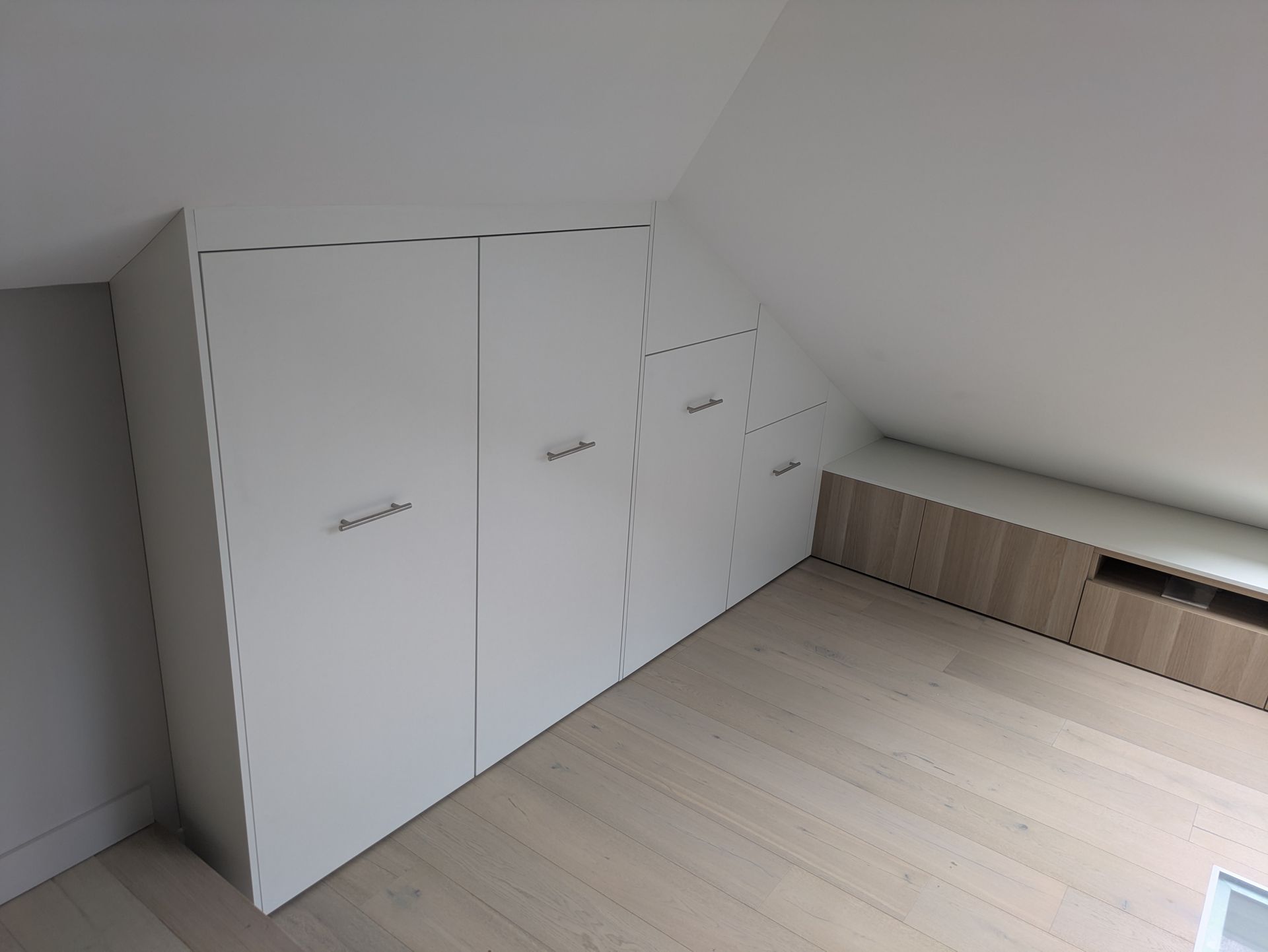 Pièce baignée de lumière avec placards en bois intégrés sous un plafond mansardé et une verrière. Un panneau de verre au sol est visible au premier plan.