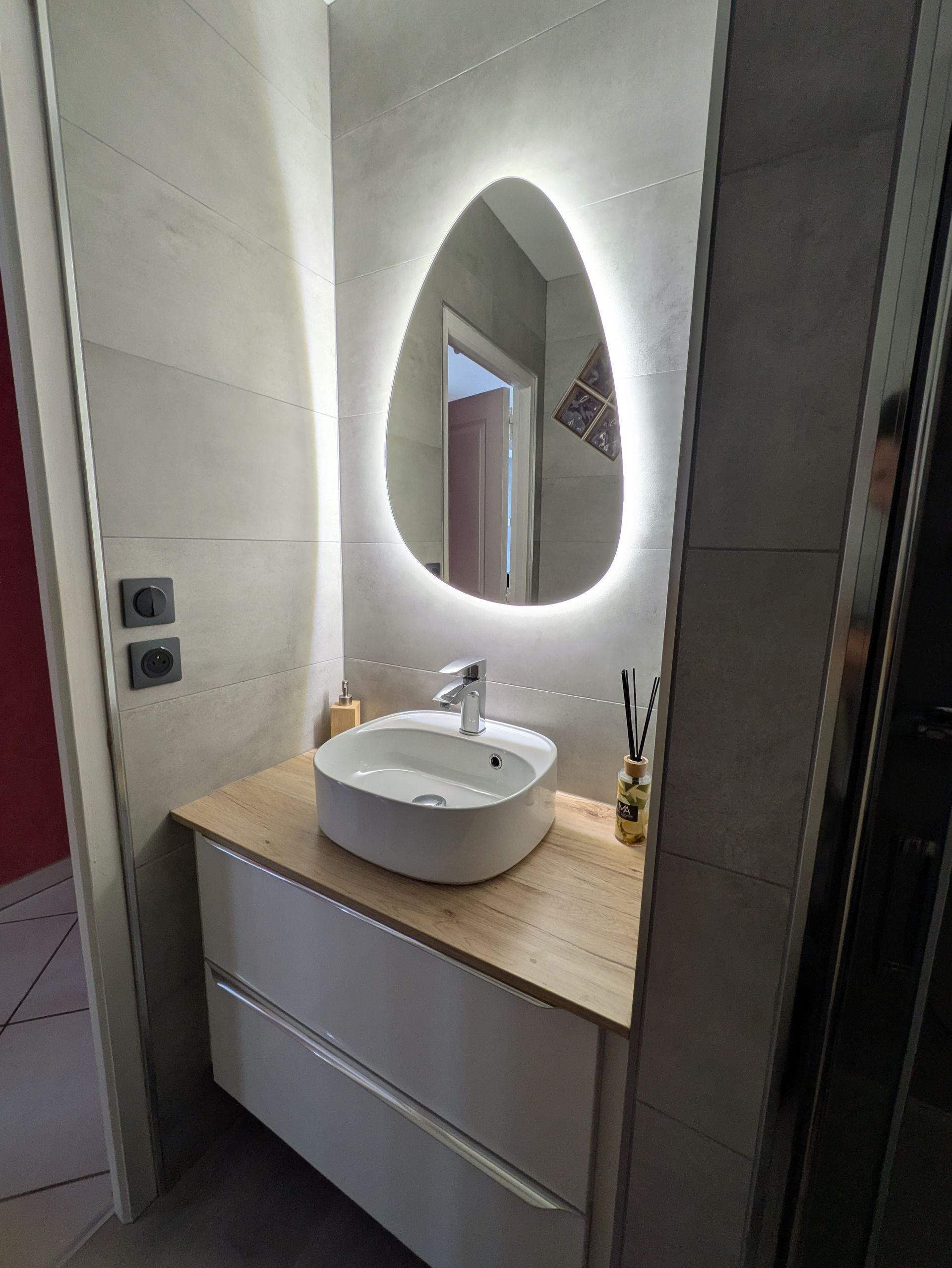 Salle de bains moderne avec miroir ovale, lavabo blanc, meuble en bois et murs carrelés gris.