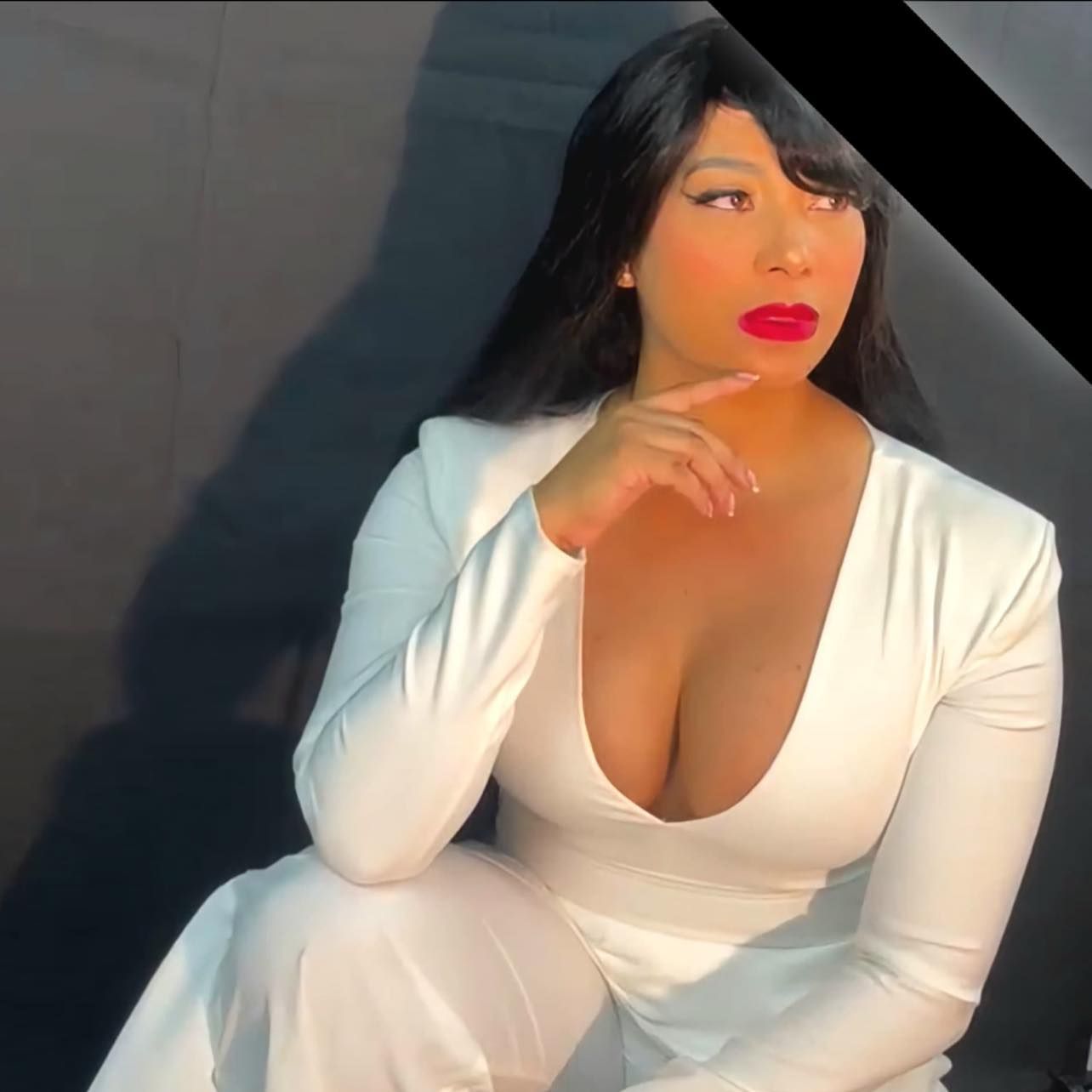 Mujer de pelo largo y negro, vestida con un mono blanco y lápiz labial rojo, con aspecto pensativo y la mano en la barbilla.