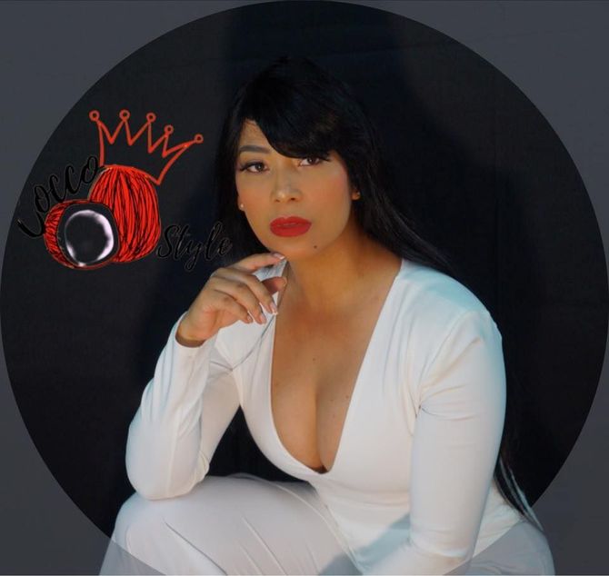 Mujer con un mono blanco, cabello oscuro con flequillo, lápiz labial rojo, posando con una mano en la barbilla, frente a un fondo oscuro con un logo.