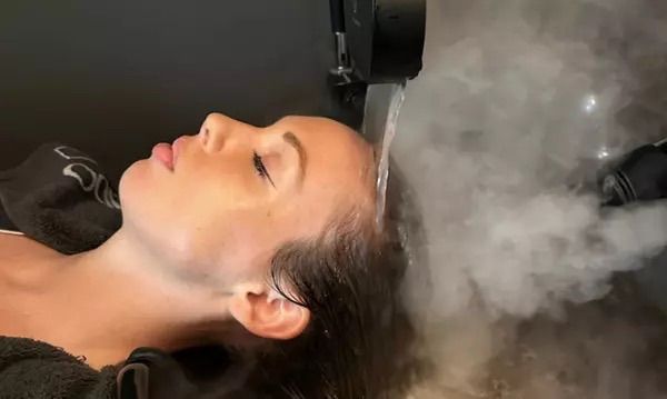 Una persona se lava el cabello en una peluquería. Un estilista con guantes negros aplica champú en el cabello de la clienta sobre un lavabo negro.