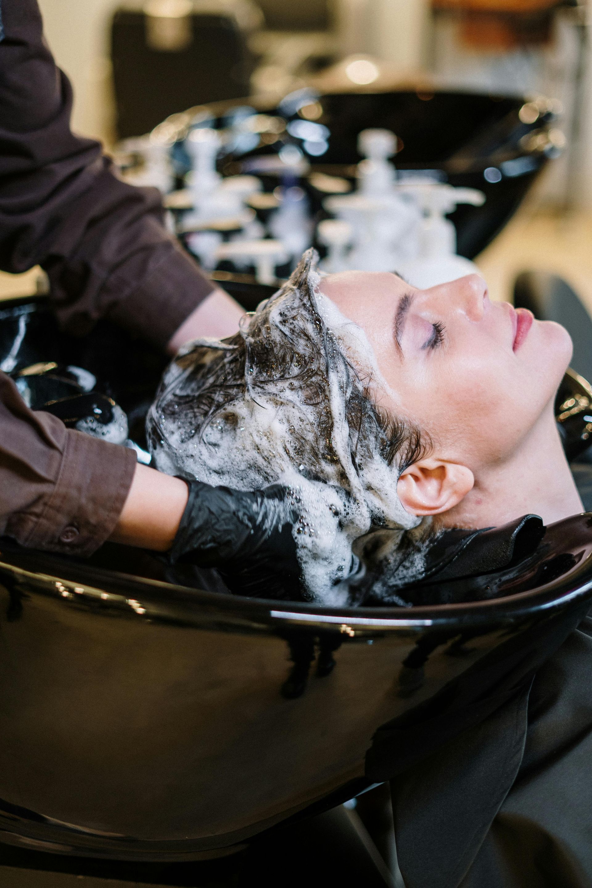 Una persona se lava el cabello en una peluquería. Un estilista con guantes negros aplica champú en el cabello de la clienta sobre un lavabo negro.