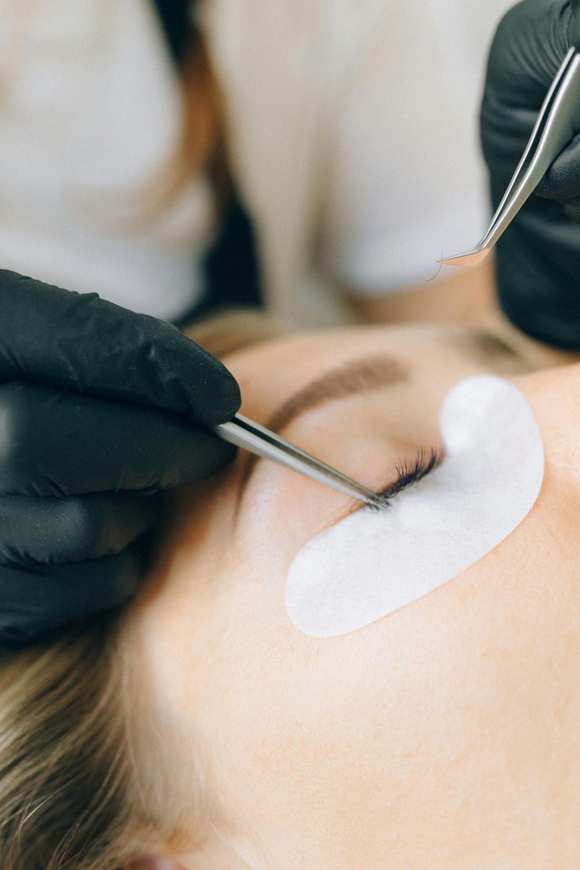 Una persona recibe extensiones de pestañas. Con guantes negros, usa pinzas para aplicar las pestañas individualmente en el párpado.