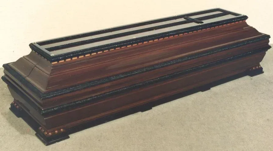 Um caixão de madeira com detalhes em preto e uma cruz na tampa, colocado sobre uma superfície de cor clara.