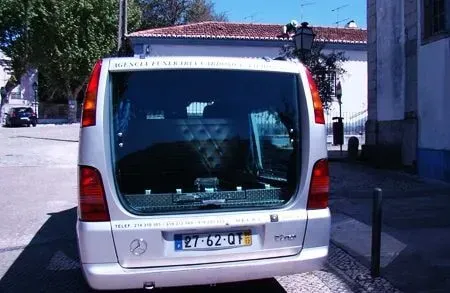Um carro funerário prateado estacionado em uma rua de paralelepípedos; ao fundo, um prédio com telhado de telha vermelha.