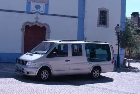 Carro funerário prateado estacionado em frente a uma igreja branca com detalhes em azul e porta vermelha.