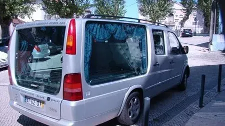 Carro funerário prateado com portas traseiras abertas numa rua, cortinas azuis visíveis no interior.