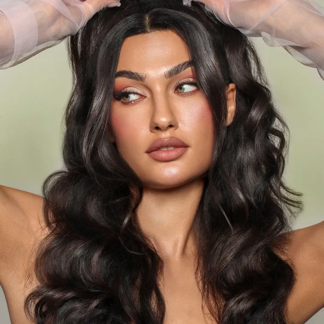 Mujer con cabello oscuro, largo y ondulado, con maquillaje ligero,