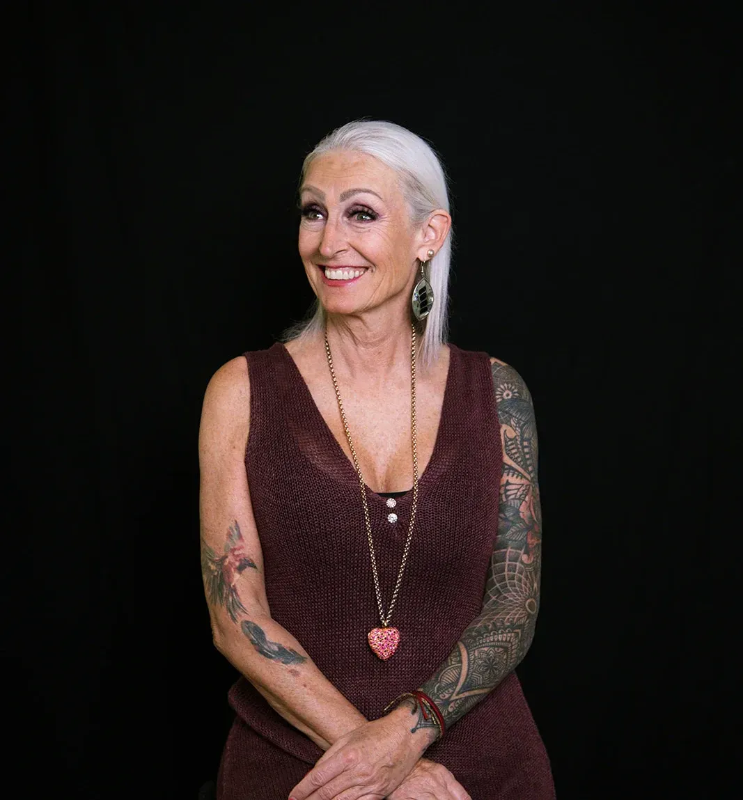 Una mujer con cabello plateado, brazos tatuados y vestido granate sonríe sobre un fondo negro.