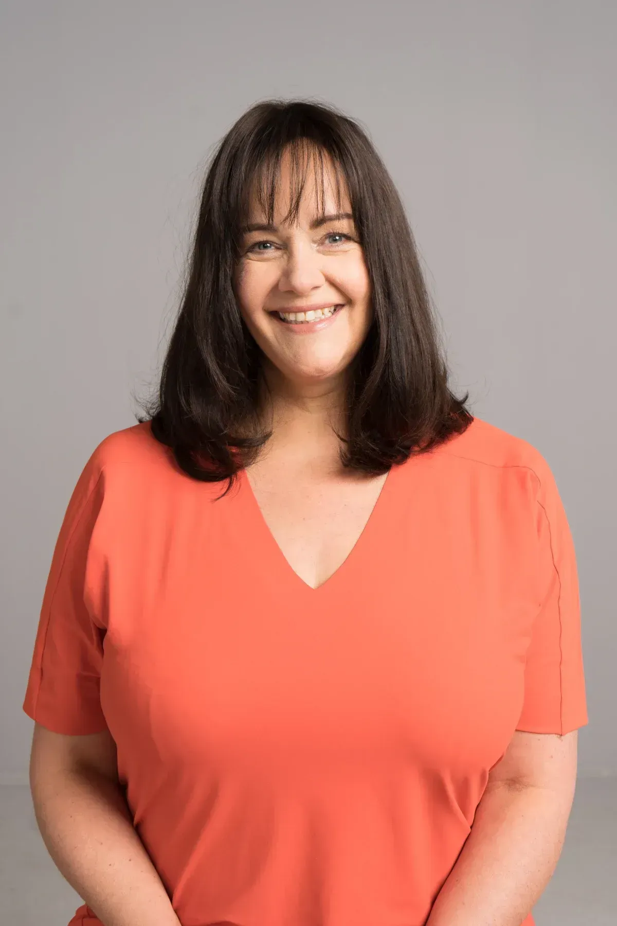 Una mujer de cabello oscuro sonríe, vistiendo una camisa naranja con cuello en V, sobre un fondo gris.