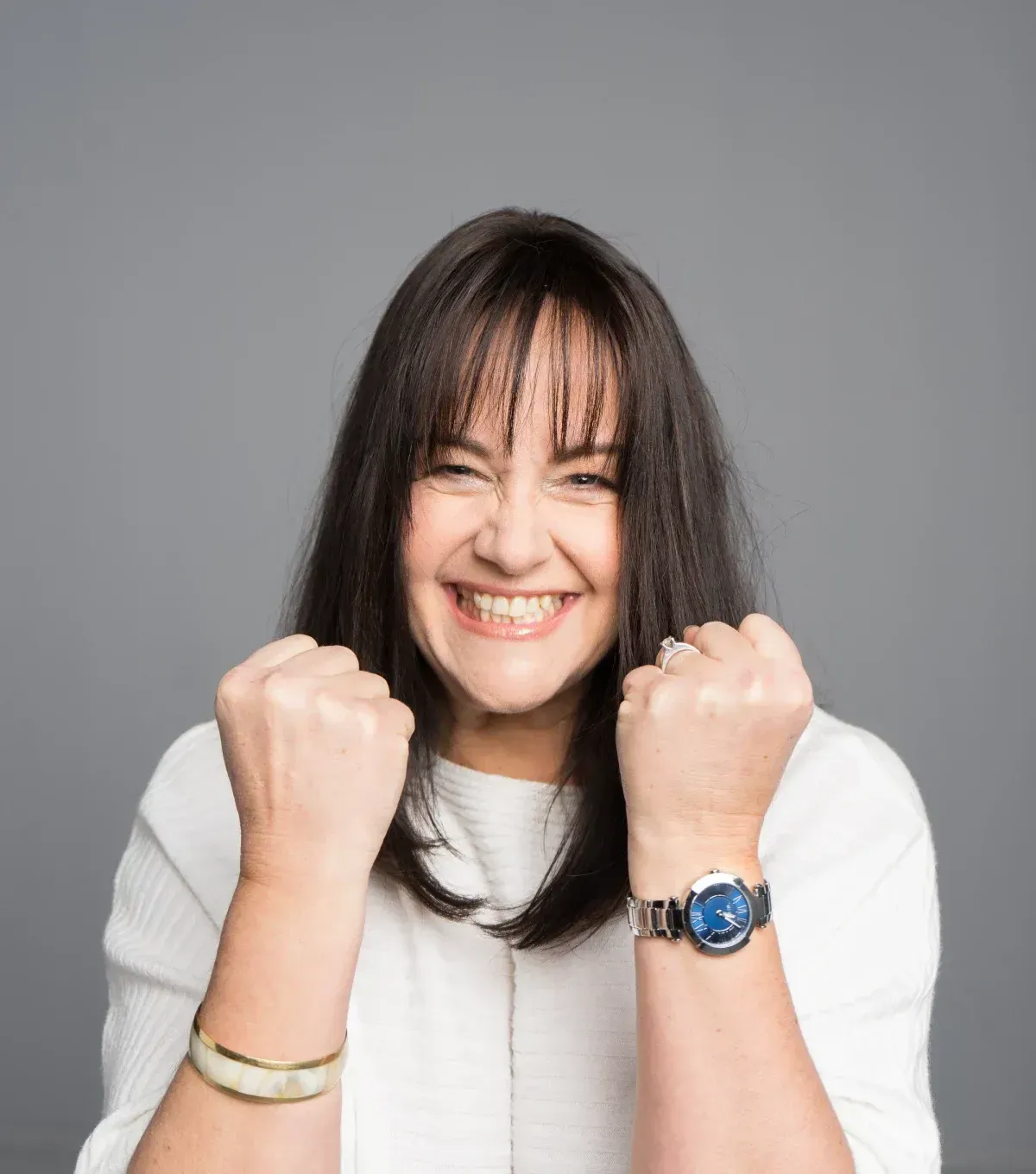Una mujer de cabello oscuro aprieta los puños y sonríe ampliamente, lleva un reloj y una pulsera, sobre un fondo gris.