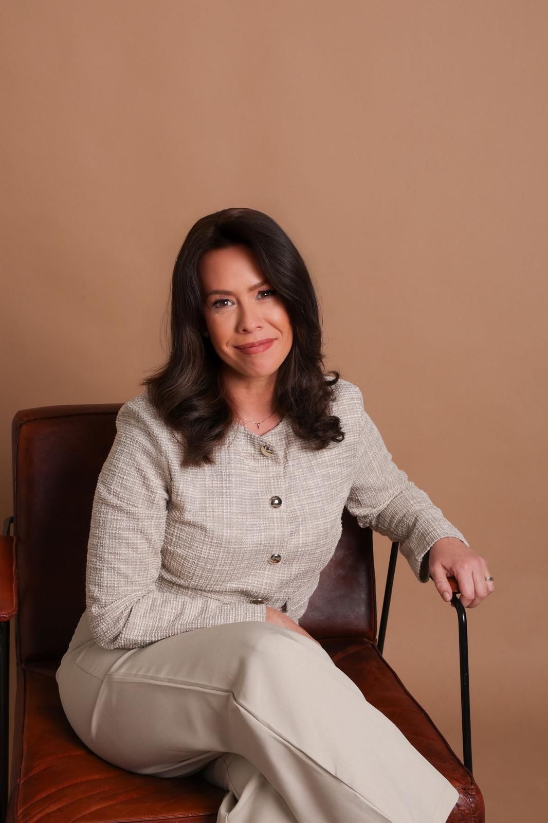 Una mujer con blazer y pantalones se sienta en una silla de cuero, sonriendo, frente a un fondo color canela.