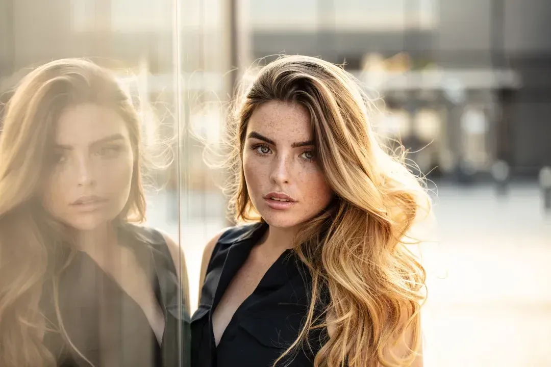Mujer con cabello rubio largo y ondulado mira a la cámara, apoyada contra una pared de vidrio, a la luz del sol.
