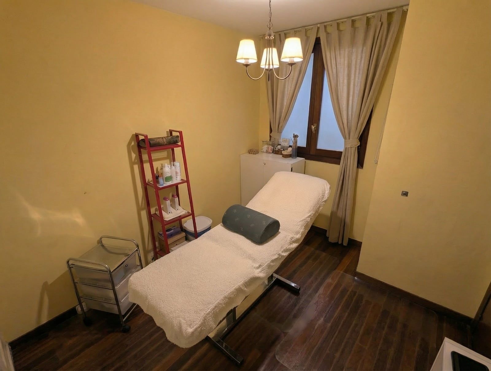 Sala de tratamientos de spa con mesa de masajes, estanterías y ventana con cortinas.
