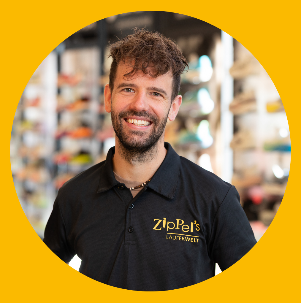 Ein Mann in einem schwarzen Zip-T-Shirt lächelt in einem Schuhgeschäft; die Logos von Adidas und Asics sind sichtbar.