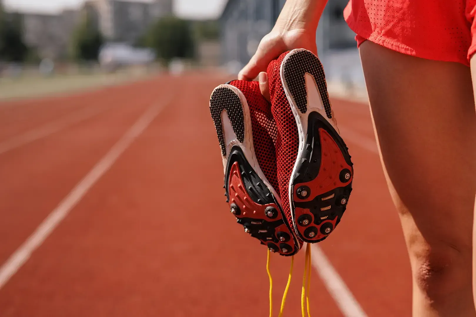 Athlet auf der Laufbahn, hält rote Laufschuhe mit gelben Schnürsenkeln, Shorts, Nahaufnahme.