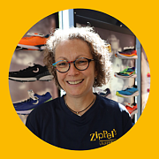 Eine Frau mit Brille lächelt in einem Schuhgeschäft, umgeben von Laufschuhen. Sie trägt ein dunkelblaues Hemd.